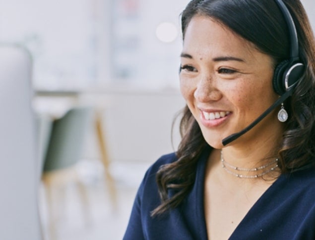 A woman with a headset smiles while working at a computer in an office setting.