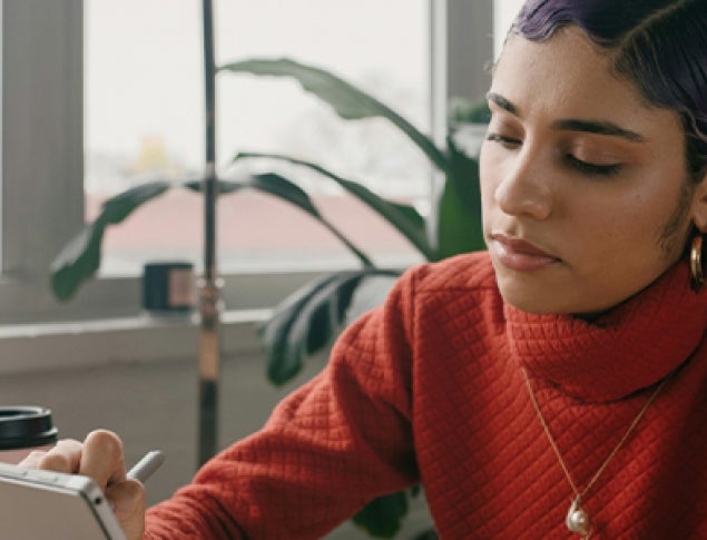 A young woman in a red sweater focuses on a tablet while seated in a bright room.