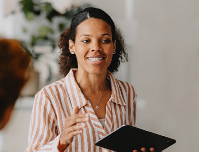 A smiling woman in a striped shirt holds a tablet while speaking to an audience.