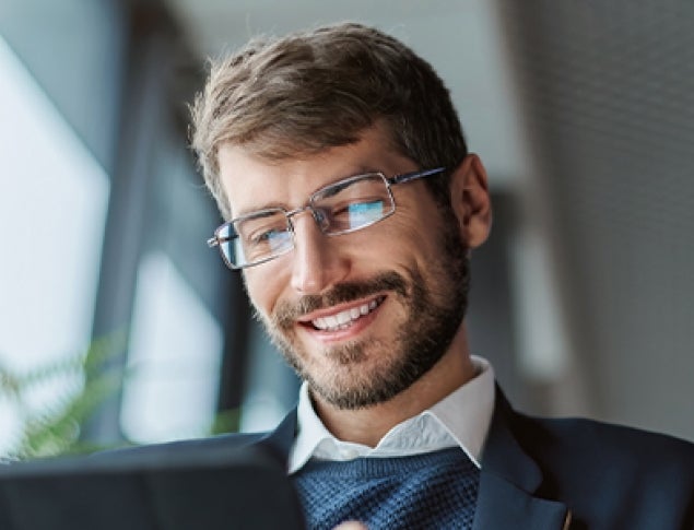 Smiling man in glasses using a tablet, sitting by a window with greenery.
