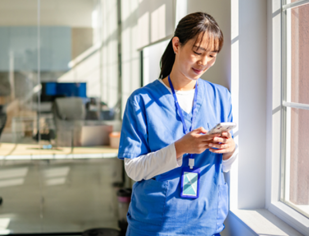 A healthcare worker in blue scrubs smiles while using a smartphone near a window.