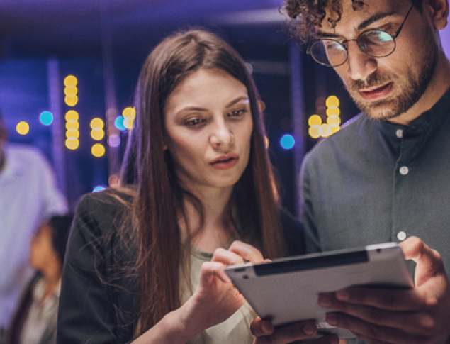 A woman and a man closely examine a tablet screen in a modern, dimly lit room.