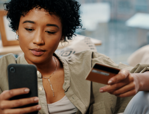 A person with curly hair holds a phone and a credit card, looking focused.