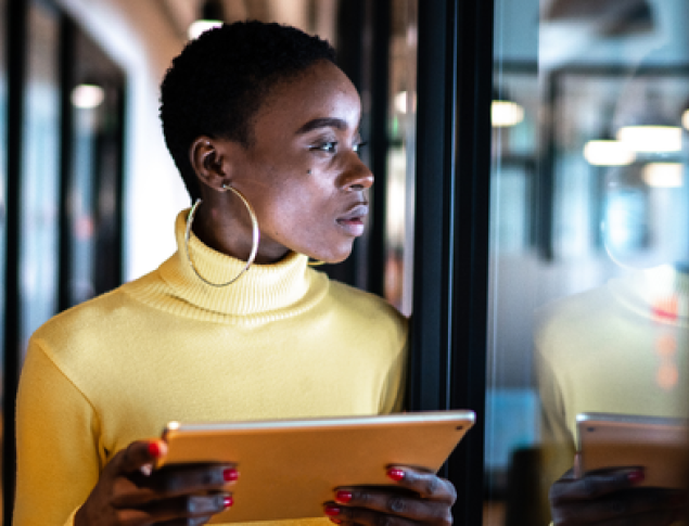 Woman in a yellow turtleneck holds a tablet, looking thoughtfully in a modern office setting.
