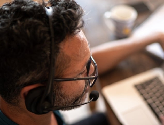 Man with curly hair and glasses wearing a headset, working on a laptop at a table.