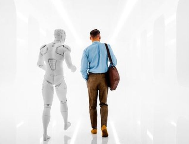 A man in a blue shirt and brown pants walks beside a translucent robotic figure in a minimalist hallway.