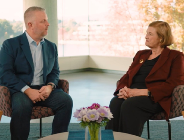 Two people sit in modern chairs, engaged in conversation, with a colorful backdrop of autumn trees.