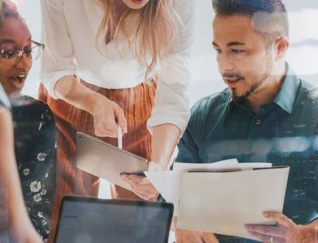 Three professionals collaborate over documents and a laptop in a bright office setting.