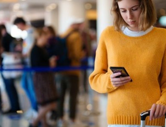 A woman in a yellow sweater checks her phone while standing near a luggage cart in an airport.