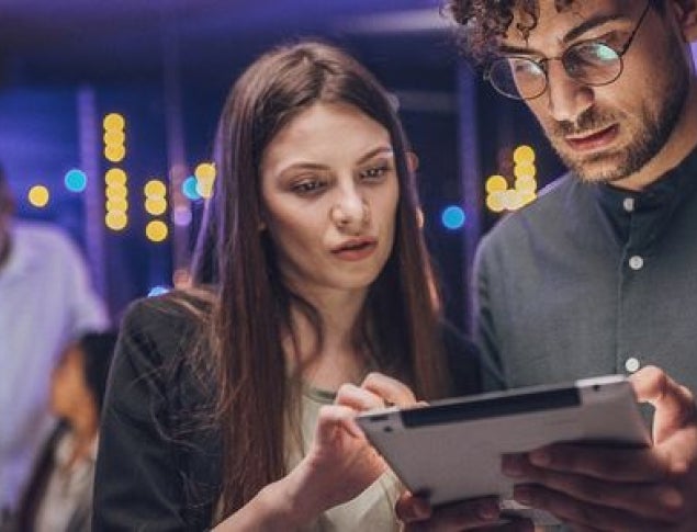 Two people closely examining a tablet, with colorful lights and another person in the background.