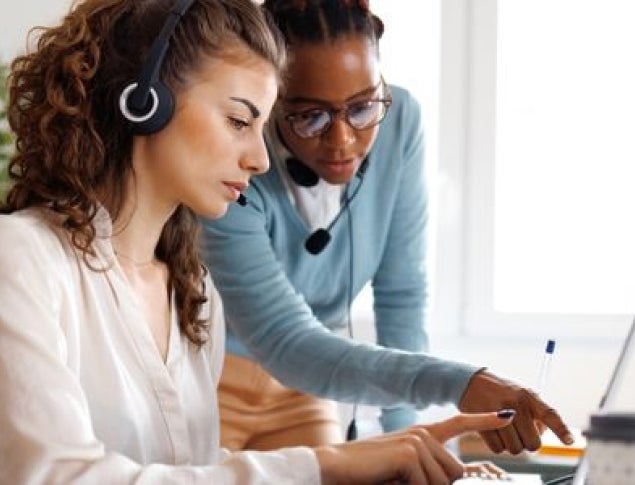 Two women collaborating at a desk; one wears a headset and the other points to a laptop screen.