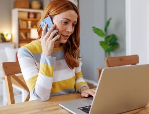 A woman with red hair sits at a wooden table, talking on her phone while working on a laptop.