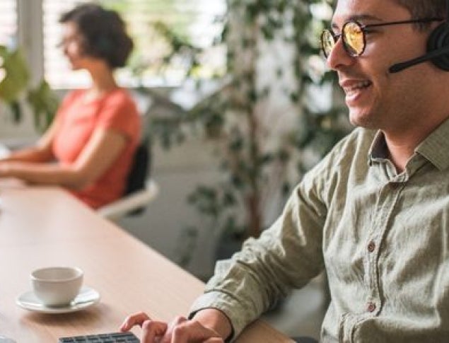 A man with glasses and a headset smiles while working at a computer, a woman sits nearby.