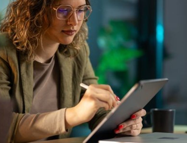 A woman with curly hair and glasses writing on a tablet at a desk with plants in the background.