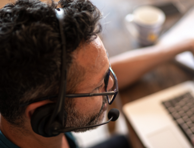 Man with curly hair and glasses wearing a headset, working on a laptop at a table.