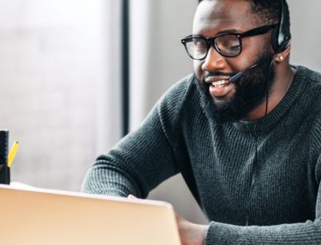 Man wearing a headset, smiling while working on a laptop in a bright office.