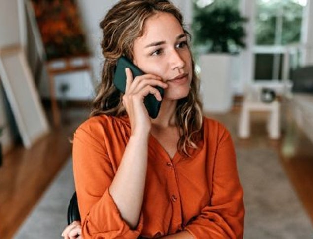 A woman with wavy hair in an orange shirt is talking on a phone in a bright room.