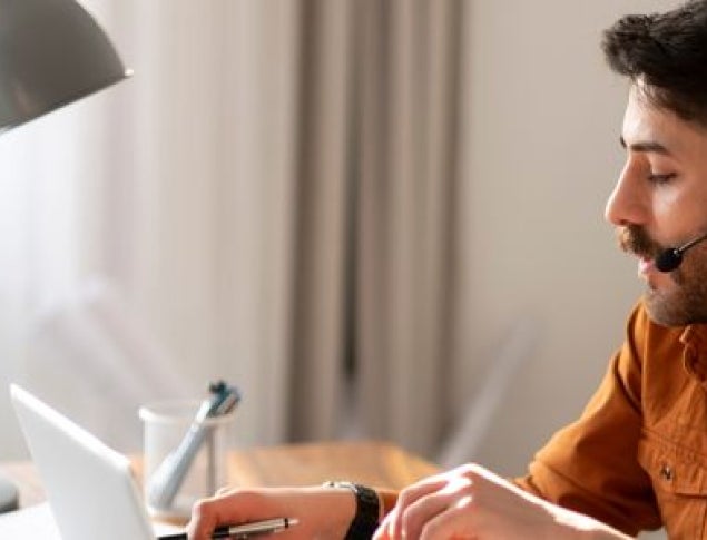 Man with headset wearing orange shirt typing notes on a computer.