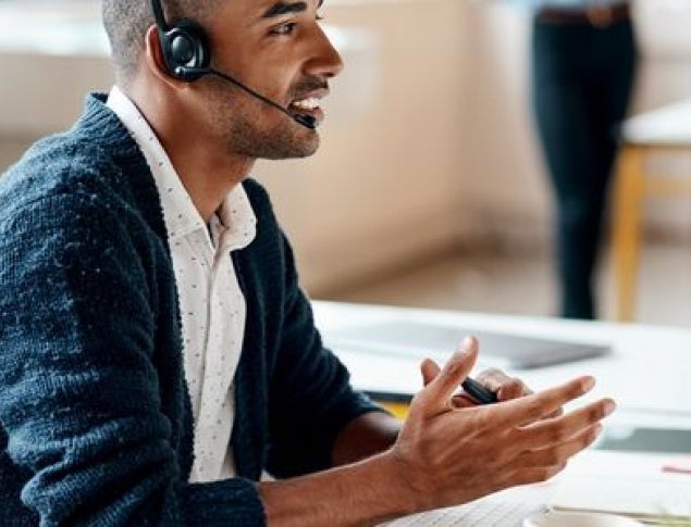Man in white patterned shirt with blue sweater wearing a headset and gesturing at desktop computer.